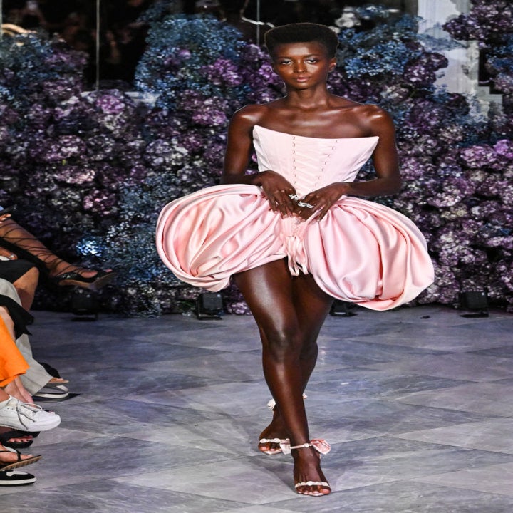 A model walks the runway in a unique, voluminous strapless dress with petal-like features, surrounded by an audience