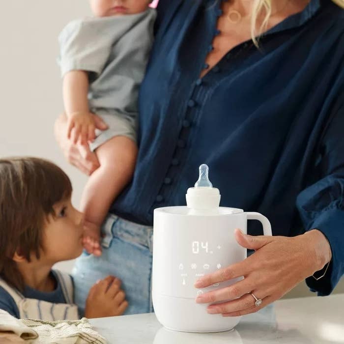 A idiosyncratic holding a baby, with a toddler beside them, uses a achromatic integer babe vessel warmer. The absorption is connected the appliance