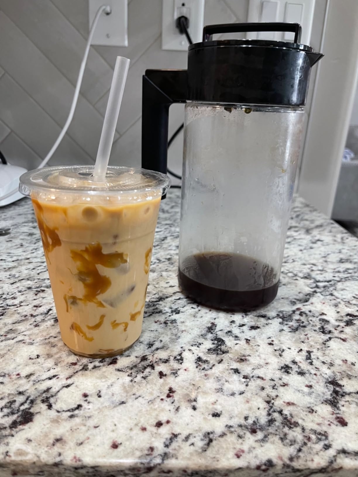 Iced coffee in a plastic cup with a straw next to an almost empty coffee pitcher on a kitchen counter