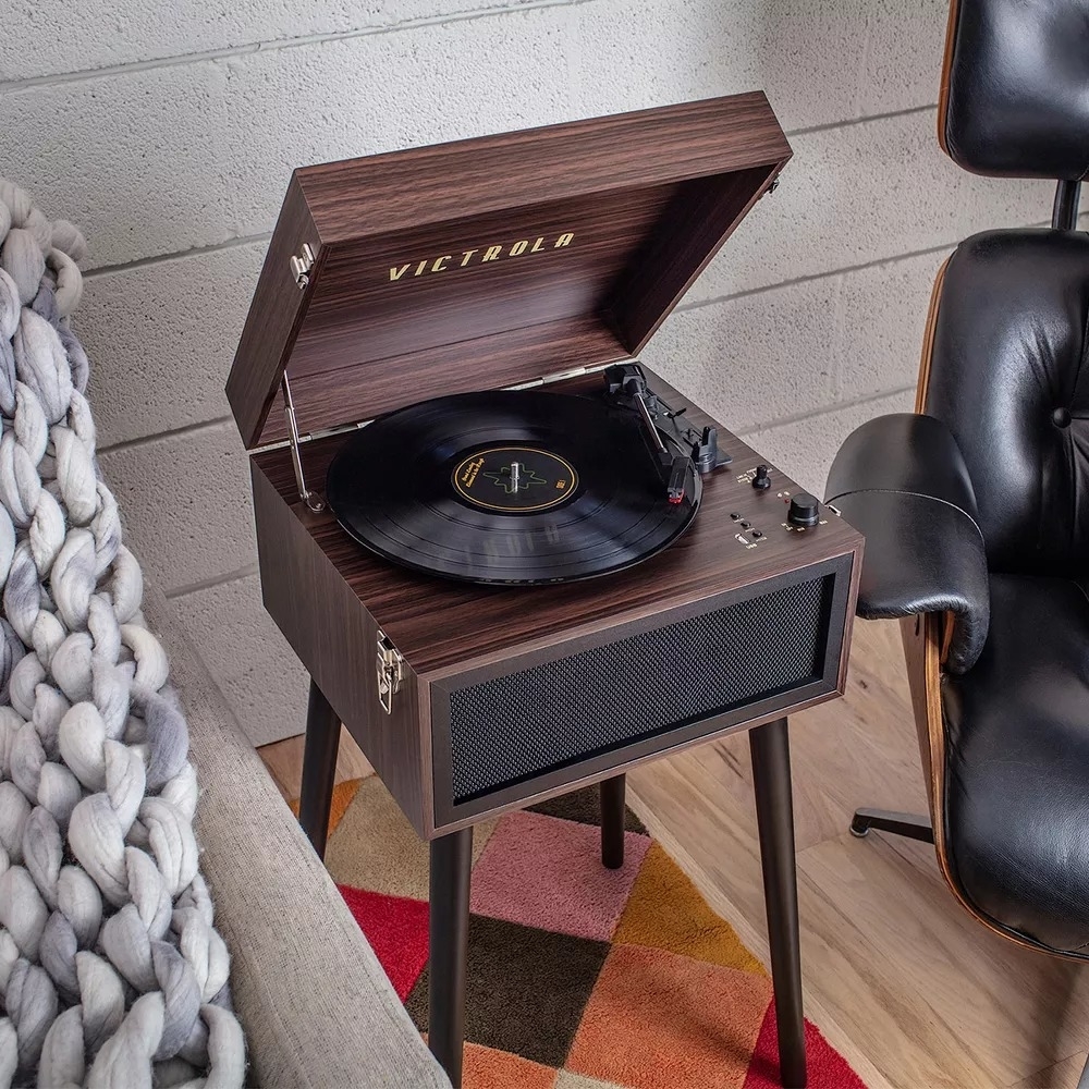 A vintage-style record player on a stand, playing a vinyl, next to a cozy chair and knit blanket, set on a patterned rug