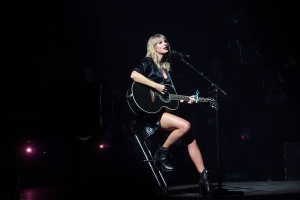 A musician plays an acoustic guitar on stage, seated on a stool, wearing a dark outfit and boots, with stage lights in the background