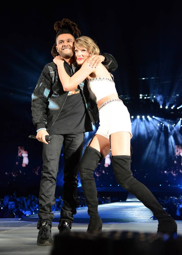 One person is in a sequin crop top and high boots, hugging another in a jacket on stage at a concert