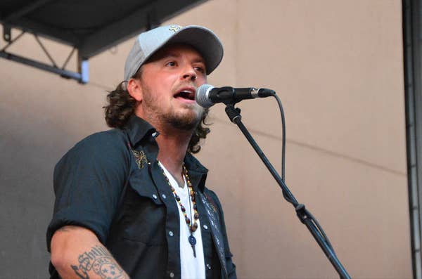 Man singing into a microphone on stage, wearing a hat and casual shirt with a tattoo visible on his arm