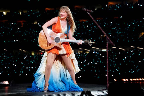 A performer in a flowing dress plays guitar on stage at a concert, surrounded by a crowd with lights