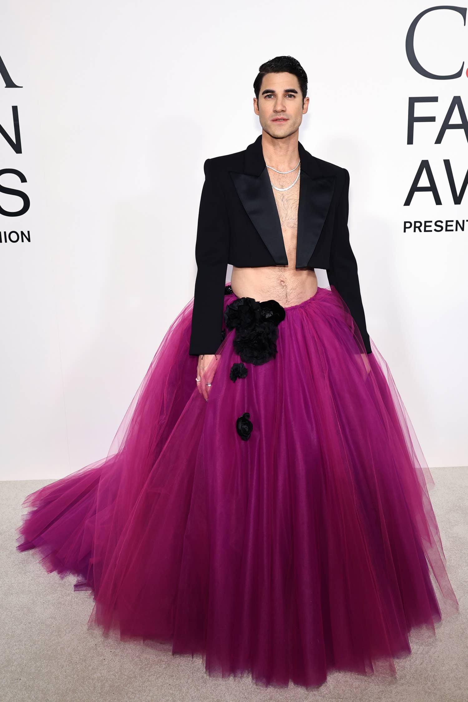 Una persona en una alfombra roja con un blazer negro corto y una voluminosa falda de tul rosa con detalles florales negros.