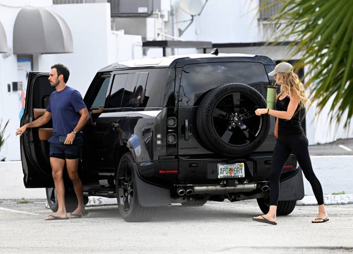 A man in a T-shirt and shorts and a woman in leggings and a cap are near a black SUV. They appear to be engaged in casual activities