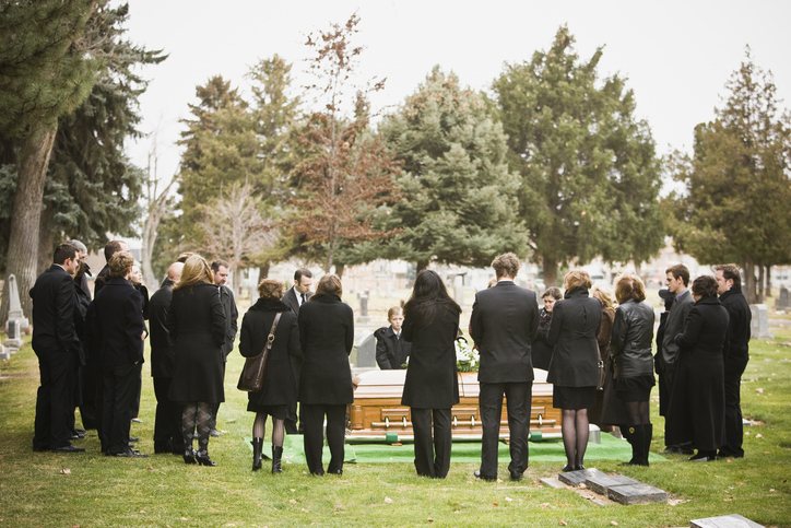 Group of people in formal black clothing gathered around a wooden coffin at an outdoor funeral service