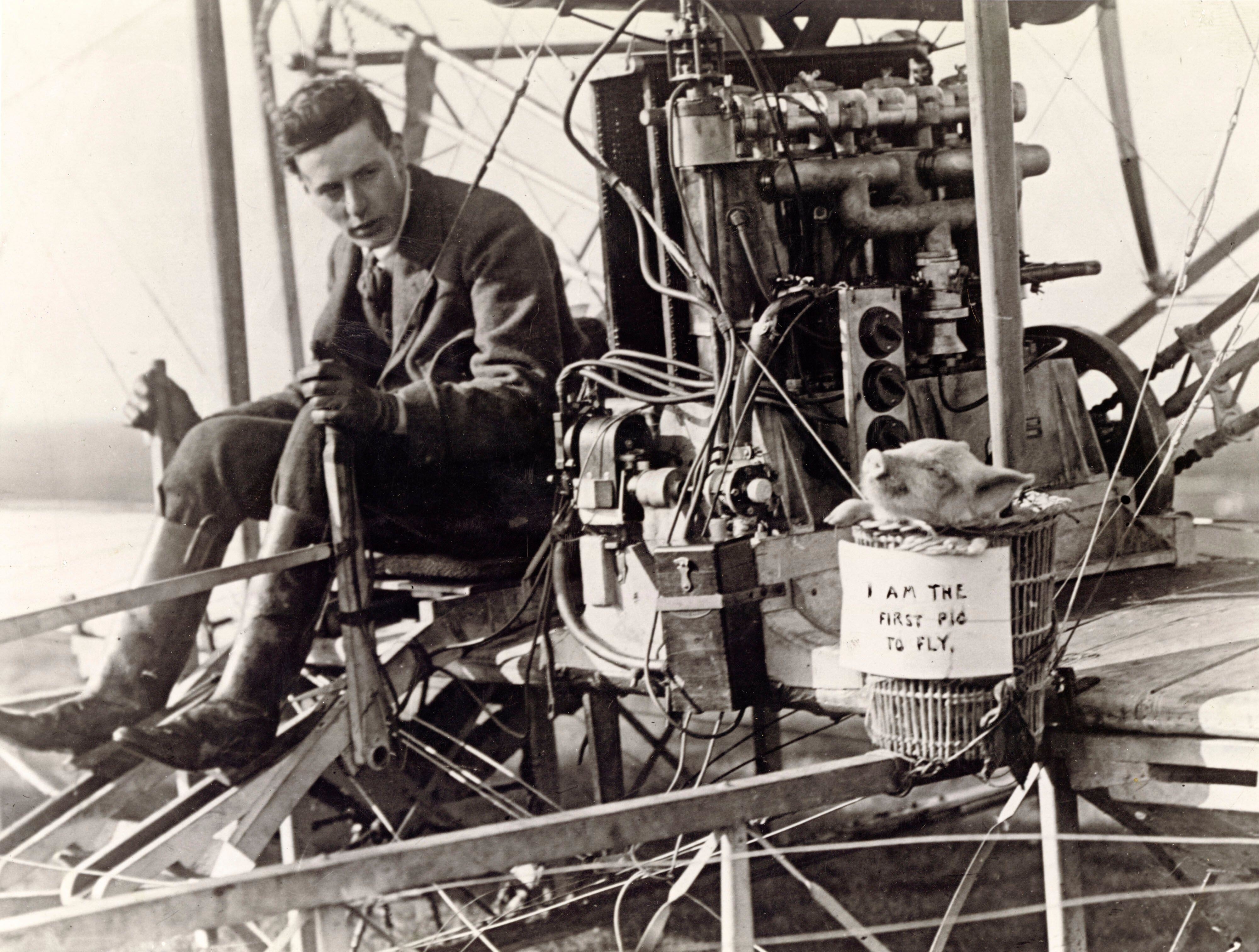 John Moore-Brabazon on his biplane with Icarus II the pig in a cage with the sign &quot;I am the first pig to fly&quot; on it