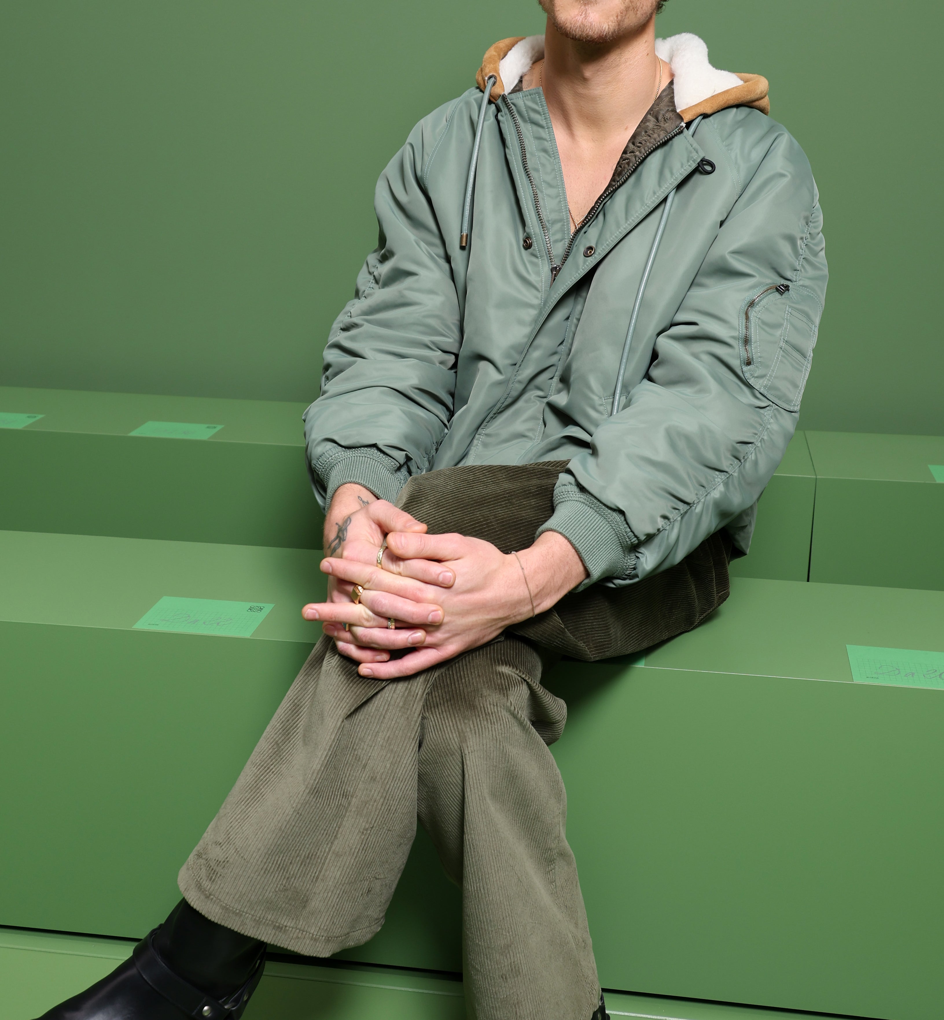 Shawn Mendes sits casually on a green bench, wearing a relaxed green bomber jacket, green pants, and black boots