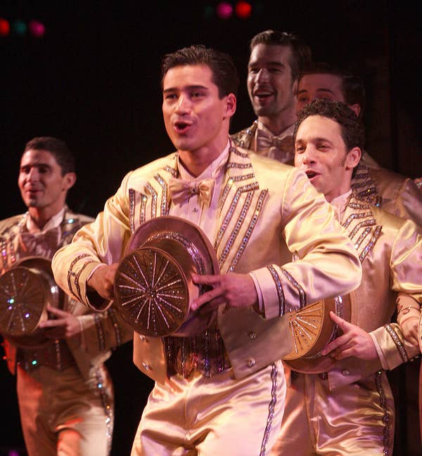 Mario front and center in group of performers in coordinated glittery formal stage costumes, holding hats