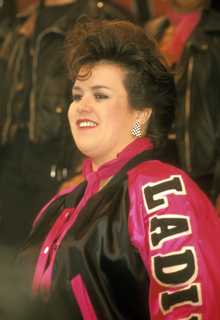 Rosie is smiling, wearing a jacket with "Ladies" on the sleeve and checkered earrings. The image is celebrity-themed
