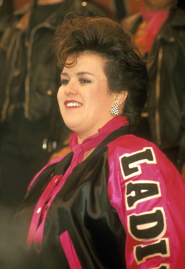 Rosie is smiling, wearing a jacket with &quot;Ladies&quot; on the sleeve and checkered earrings. The image is celebrity-themed