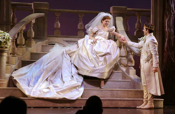 Carly in an elaborate ballgown and veil walking down stairs, holding hands with a prince