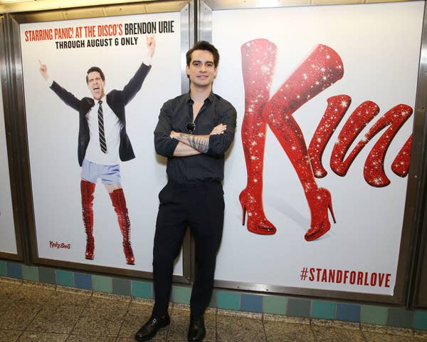 Brendan standing in front of a &quot;Kinky Boots&quot; poster featuring himself in sparkly thigh high boots. The poster highlights the show&#x27;s dates and hashtag #StandForLove