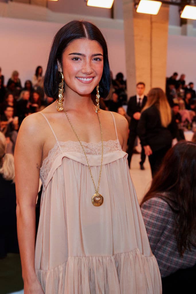 Charli wearing a loose, sleeveless dress with gold jewelry at a fashion event. Crowd visible in the background