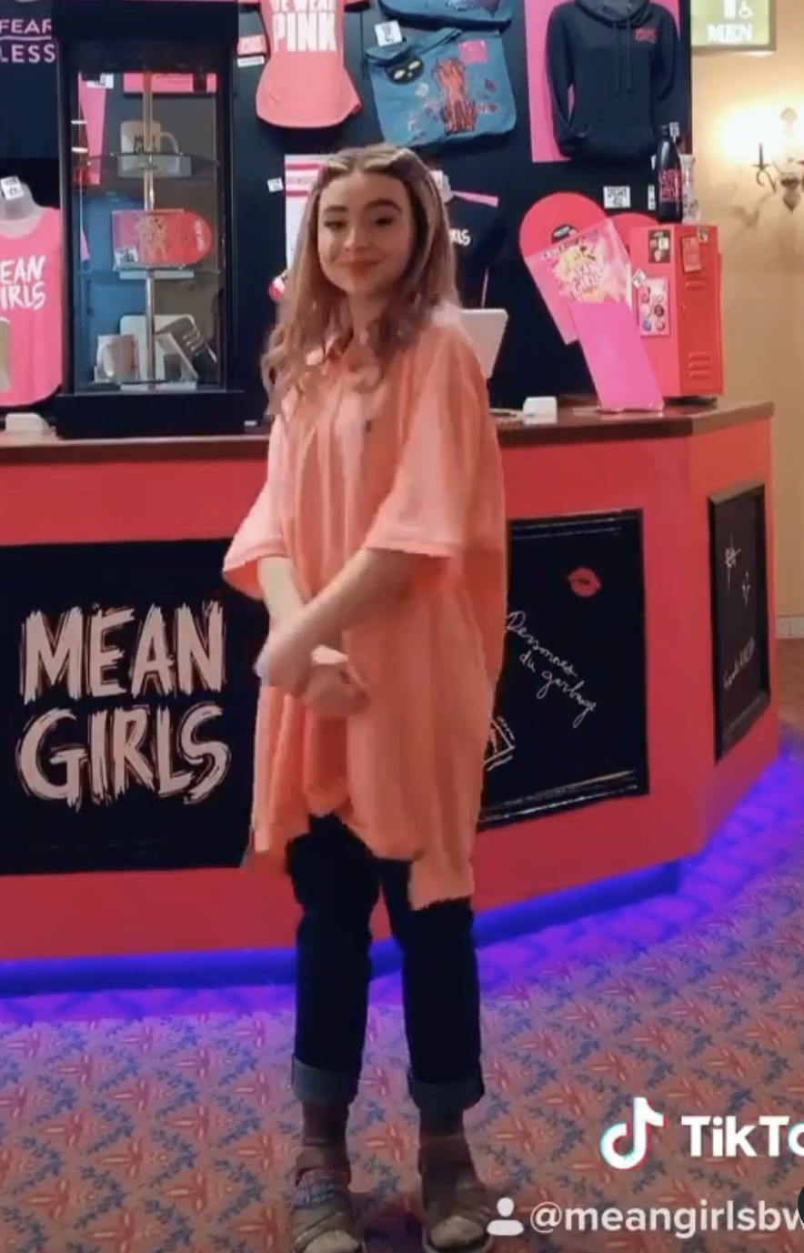 Sabrina stands in front of a "Mean Girls" merchandise display, wearing a casual oversized shirt and dark pants