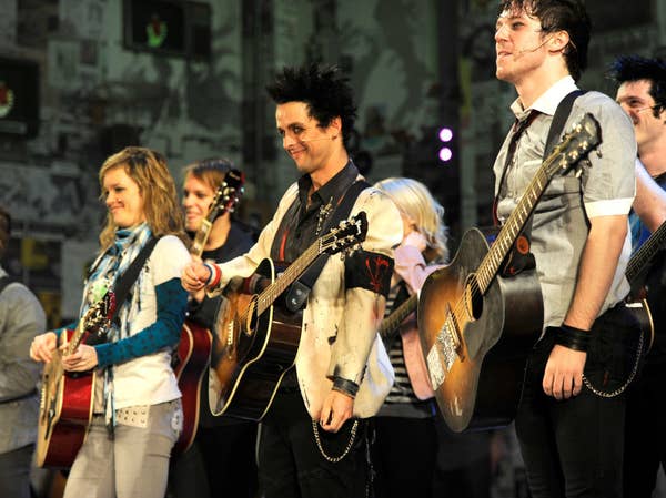 People stand with guitars on stage, including Billie Joe Armstrong. They&#x27;re smiling and dressed casually