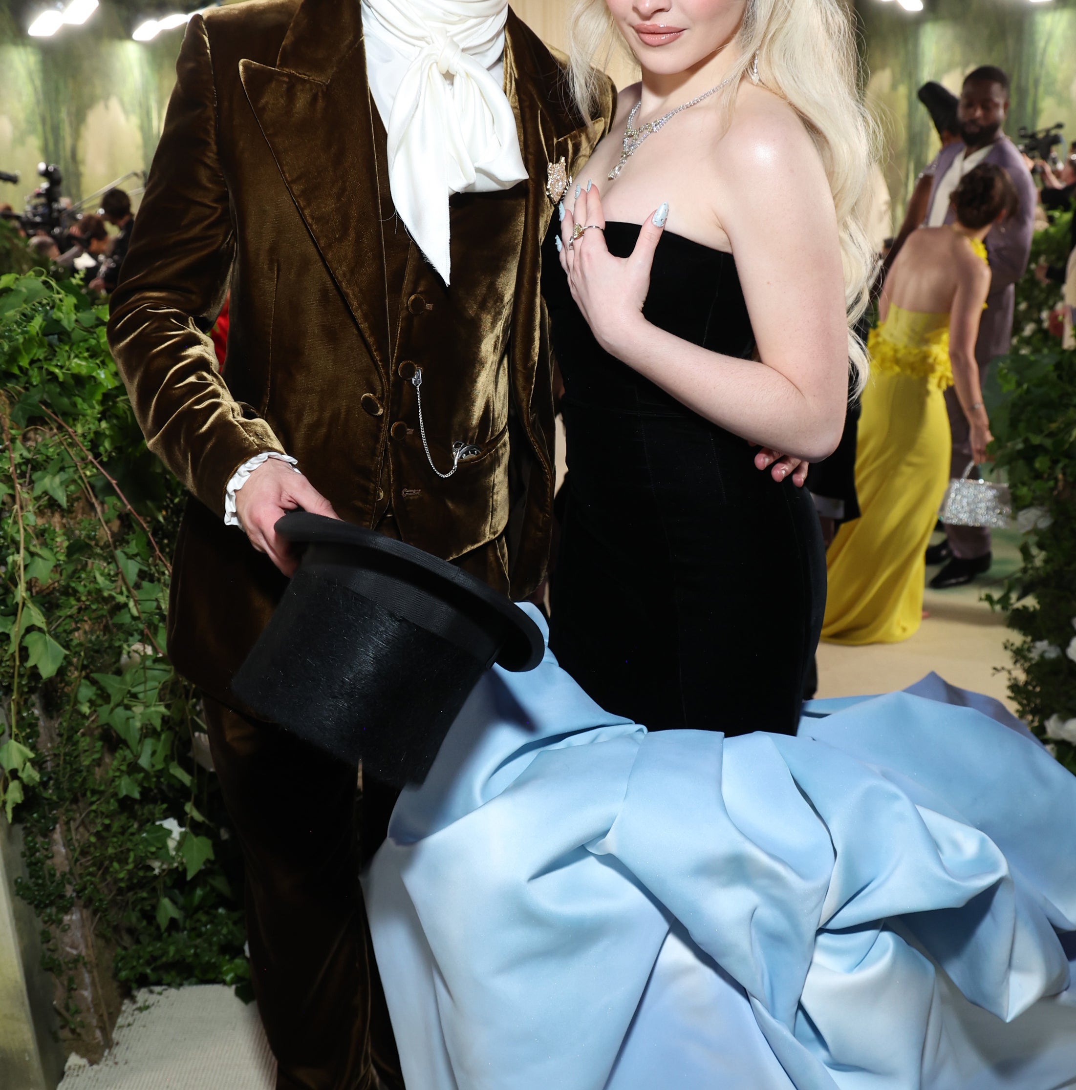 Barry Keoghan in a brown suit with a white scarf holds a top hat. Sabrina Carpenter wears a strapless black gown with a long light blue train on a formal event carpet