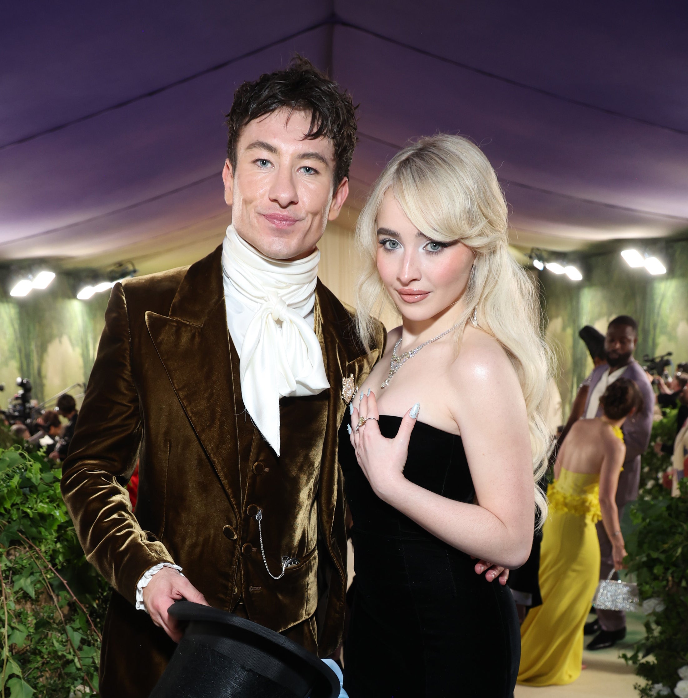 Barry Keoghan in a brown suit with a white scarf holds a top hat. Sabrina Carpenter wears a strapless black gown with a long light blue train on a formal event carpet