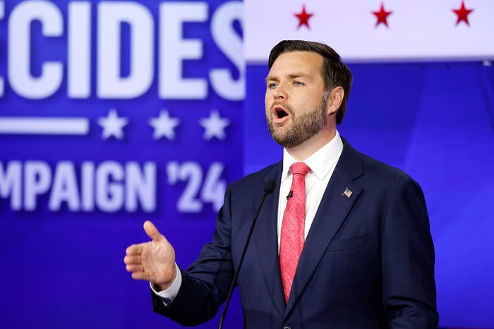J.D. in a suit and tie speaks passionately at a campaign event with the theme "Decides" and "Campaign '24" in the background
