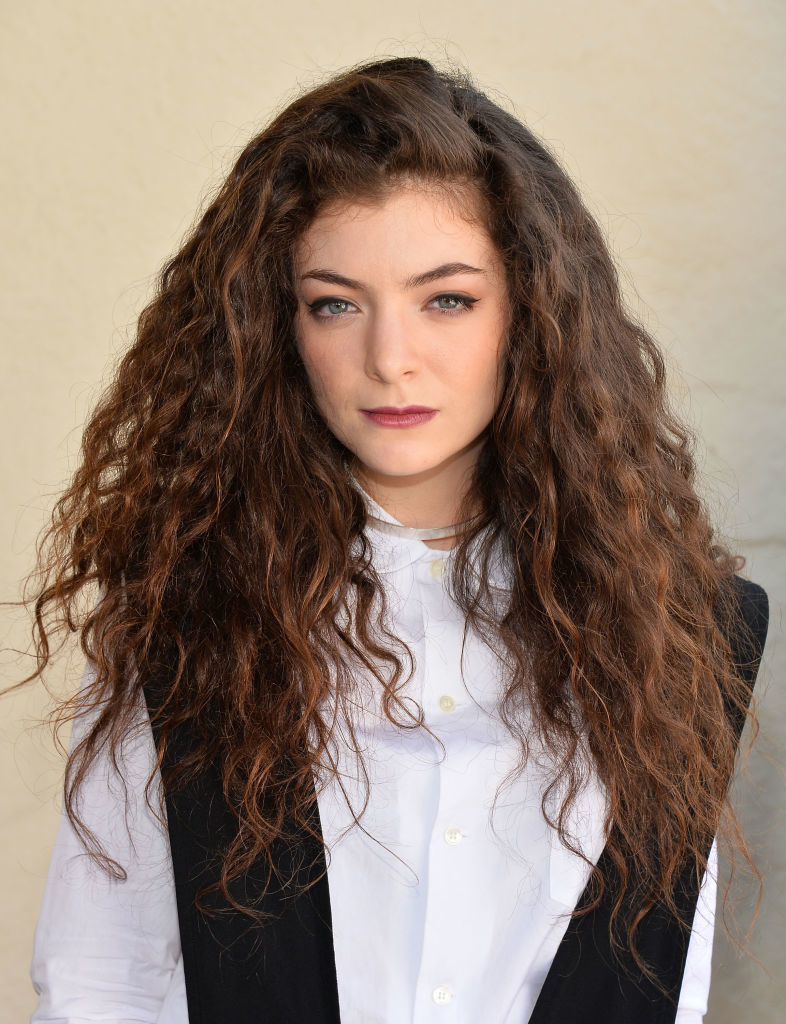 A person with long, curly hair wearing a white shirt and dark vest, looking directly at the camera