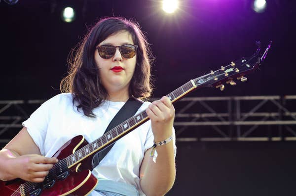 A person plays an electric guitar on stage wearing sunglasses and a white t-shirt under bright lights