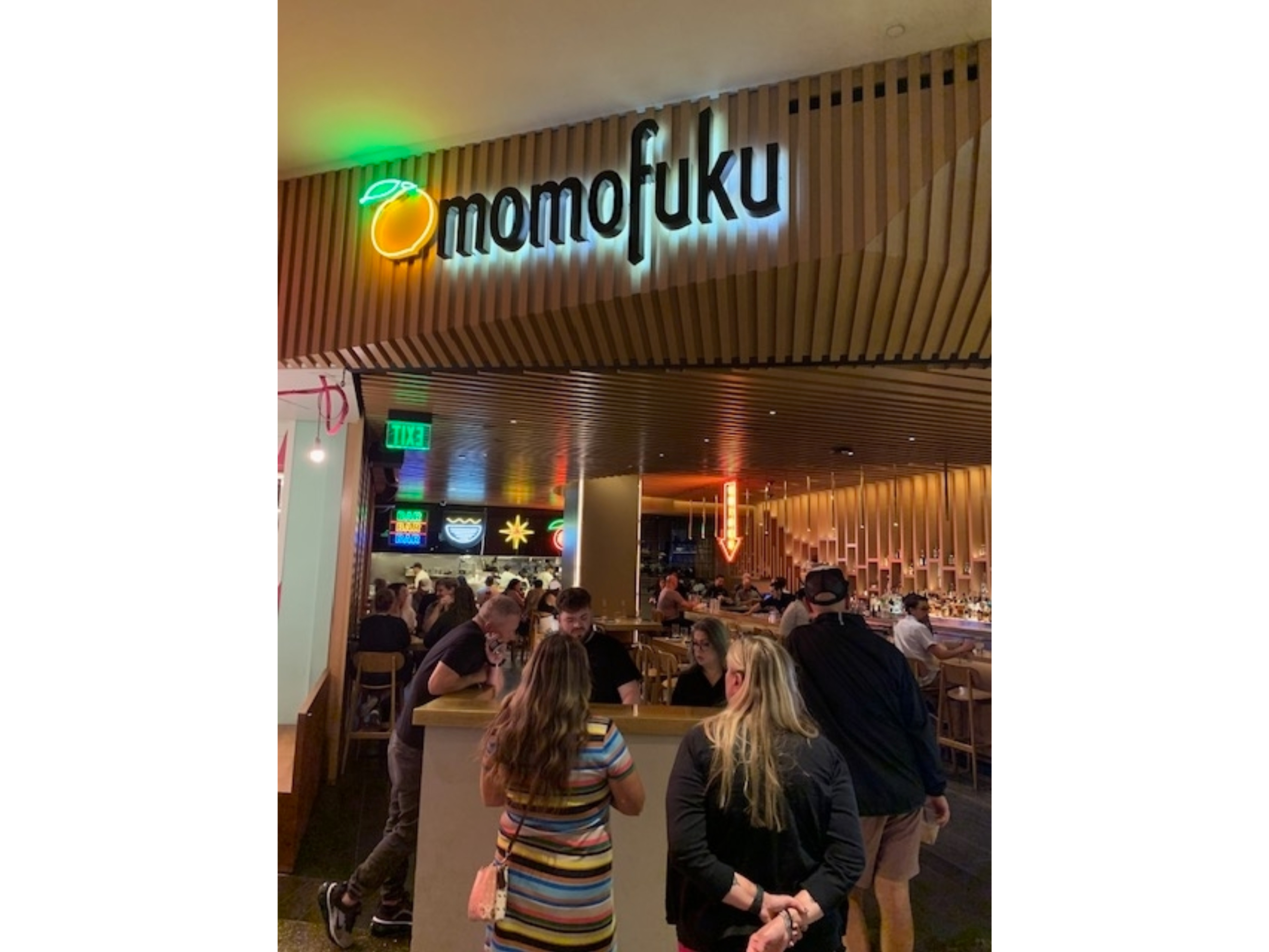 People at a Momofuku restaurant counter, with bright, modern decor and a visible neon sign