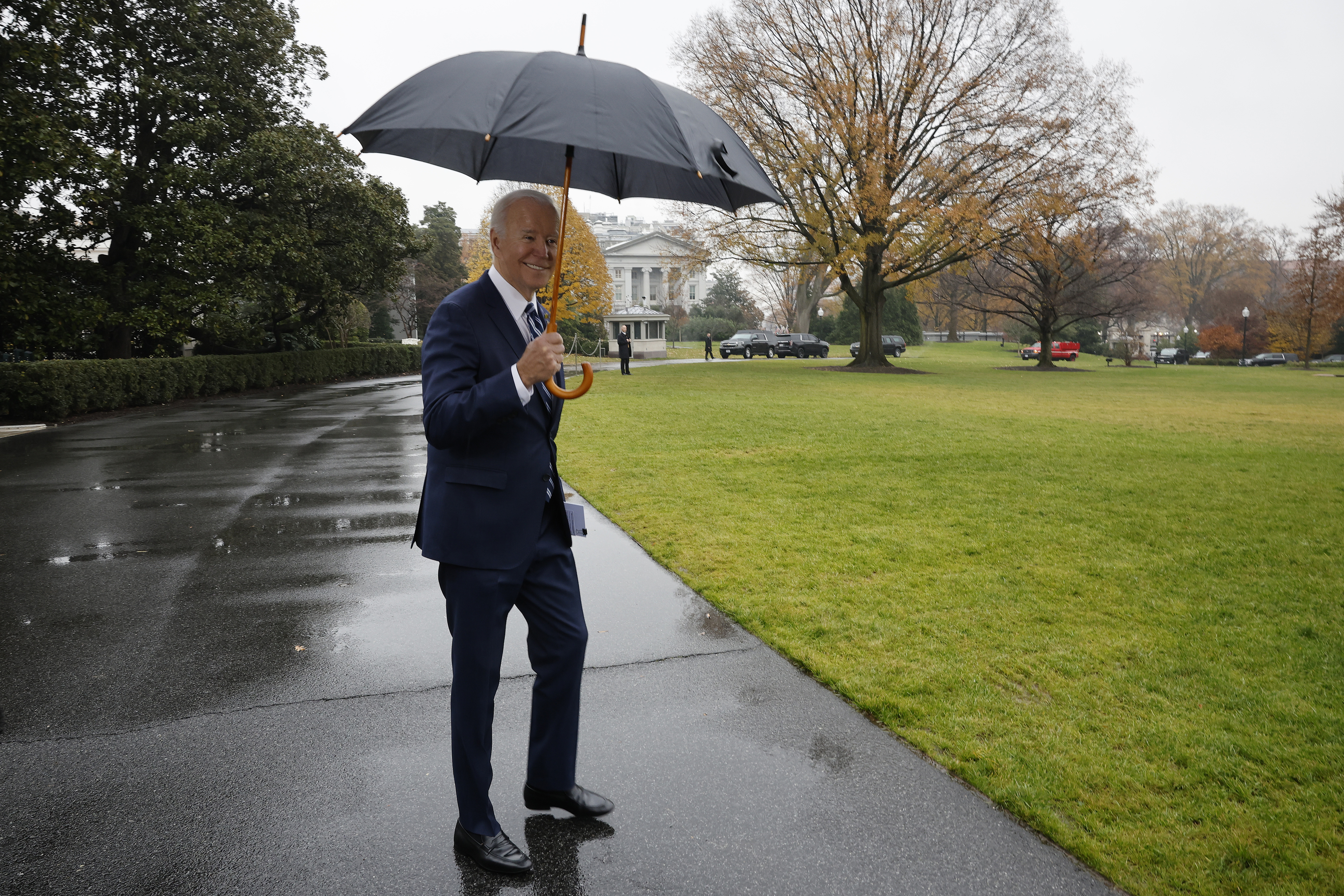 A person in a suit holds an umbrella while walking on a wet path in front of a large house with trees in the background