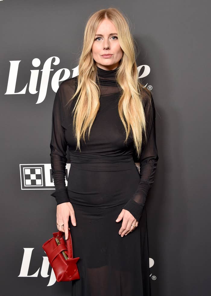 A woman with long hair in a sheer long-sleeved black dress holds a red clutch on a red carpet