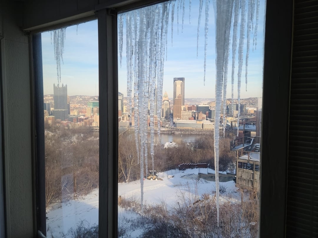 View of a snowy cityscape from a window with large icicles hanging down; Pittsburgh skyline in the background
