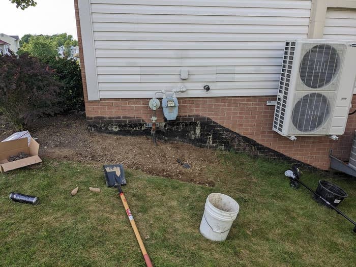 Exterior wall with an air conditioning unit, exposed dirt patch, and surrounding tools like a shovel and bucket; appears to be home maintenance work