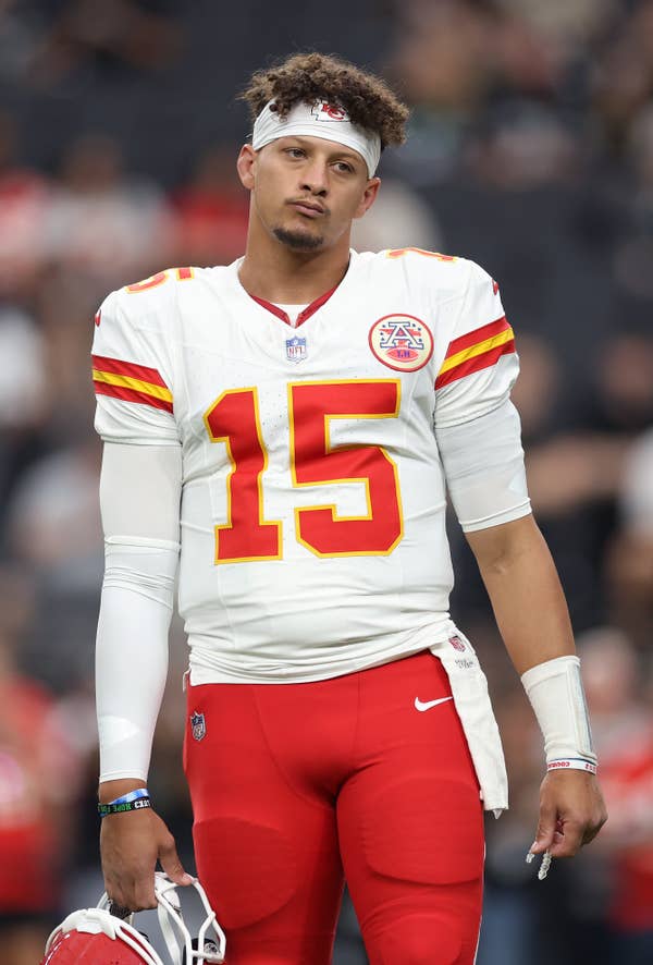 A football player in a Kansas City Chiefs uniform stands on the field, holding a helmet