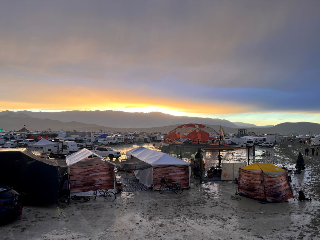 Festival tents and structures in a muddy field at sunset, with mountains in the background