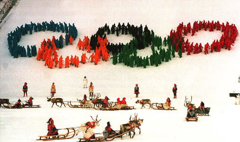 People in colorful outfits form Olympic rings on snowy ground, with reindeer and sleds in the foreground