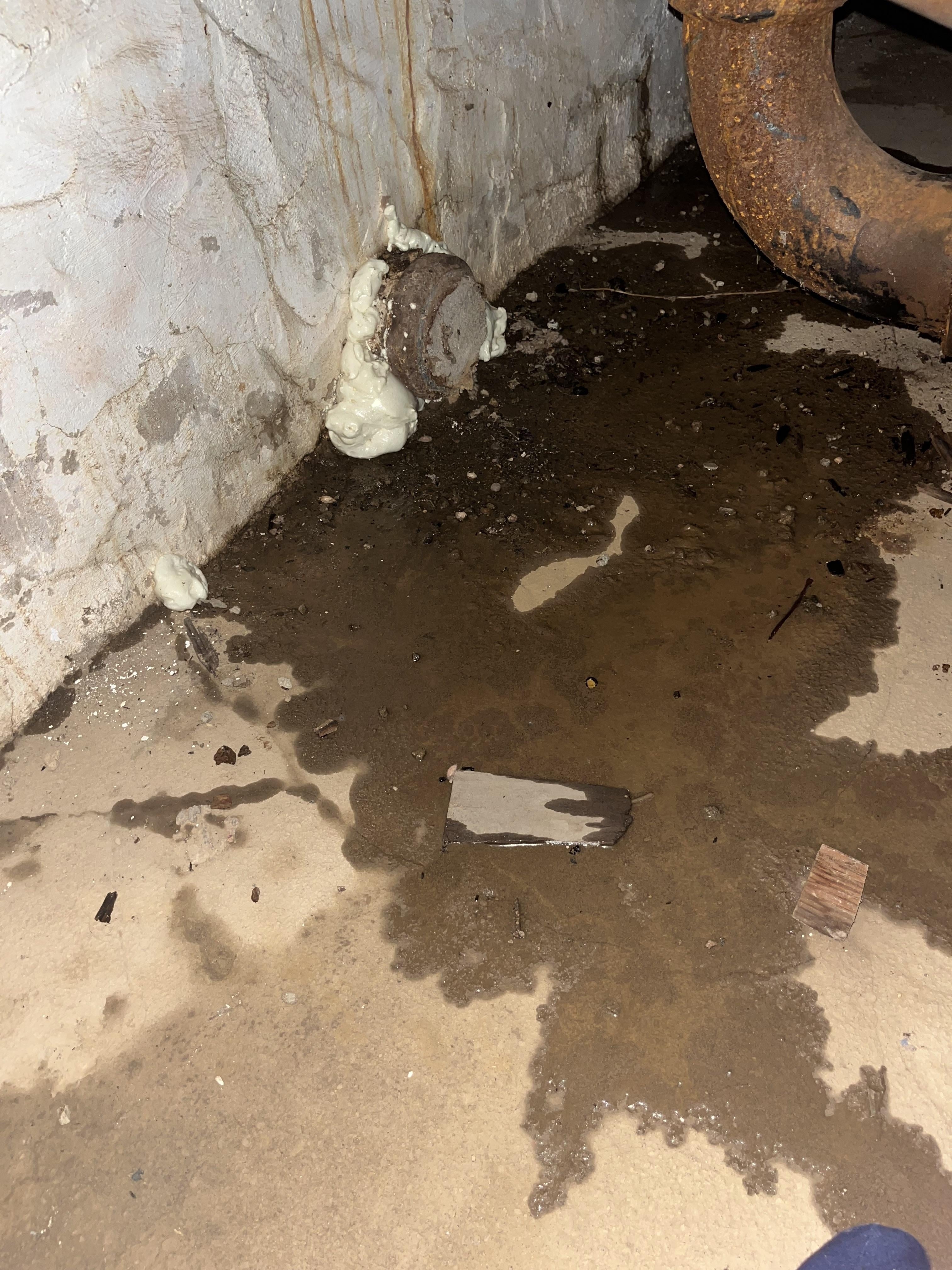 Basement corner with water seepage and dampness, showing exposed pipe and surrounding debris on the floor