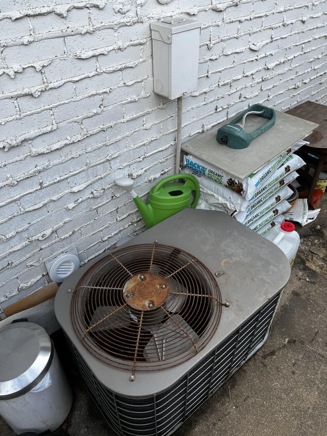 An outdoor air conditioning unit near a white brick wall is surrounded by gardening supplies, including a green watering can and stacked soil bags