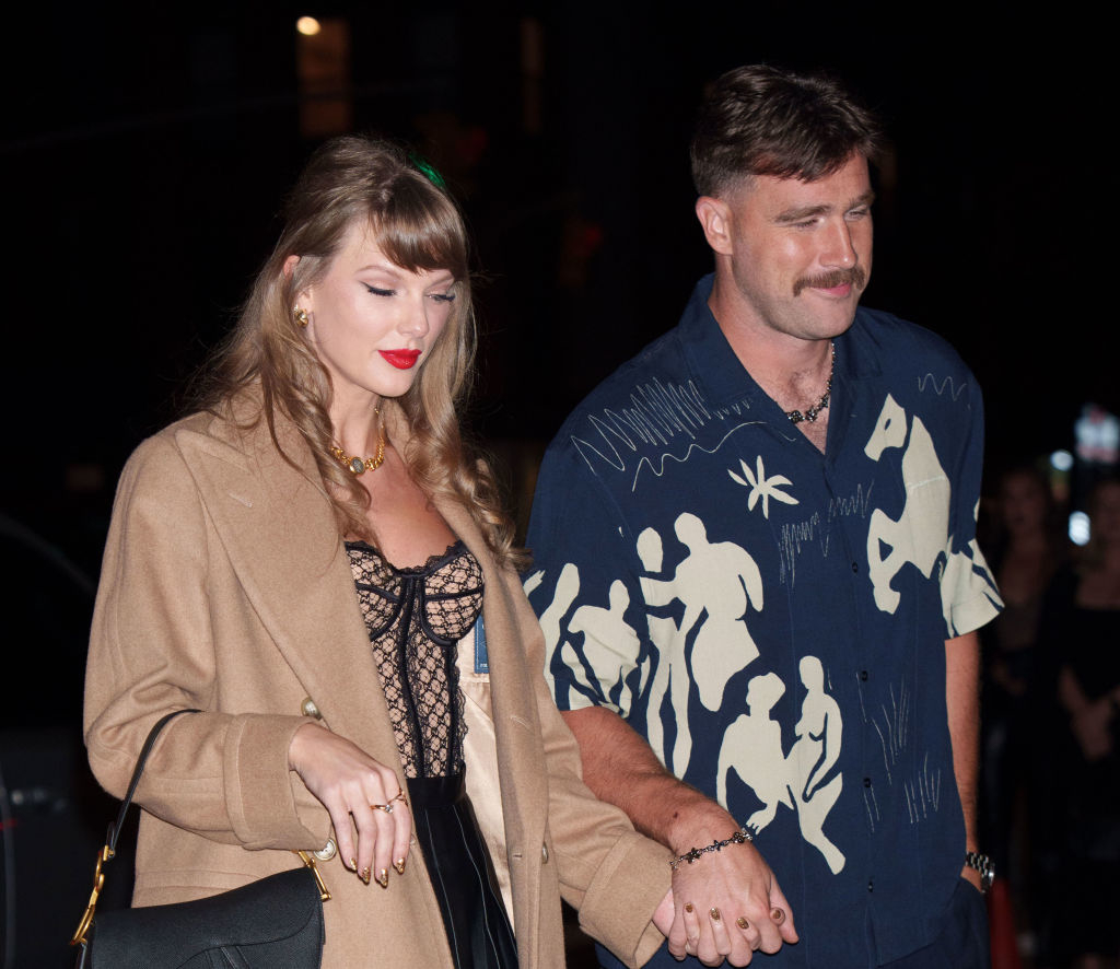 A woman in a patterned top and jacket walks hand-in-hand with a man wearing a graphic print shirt. They are outdoors at night