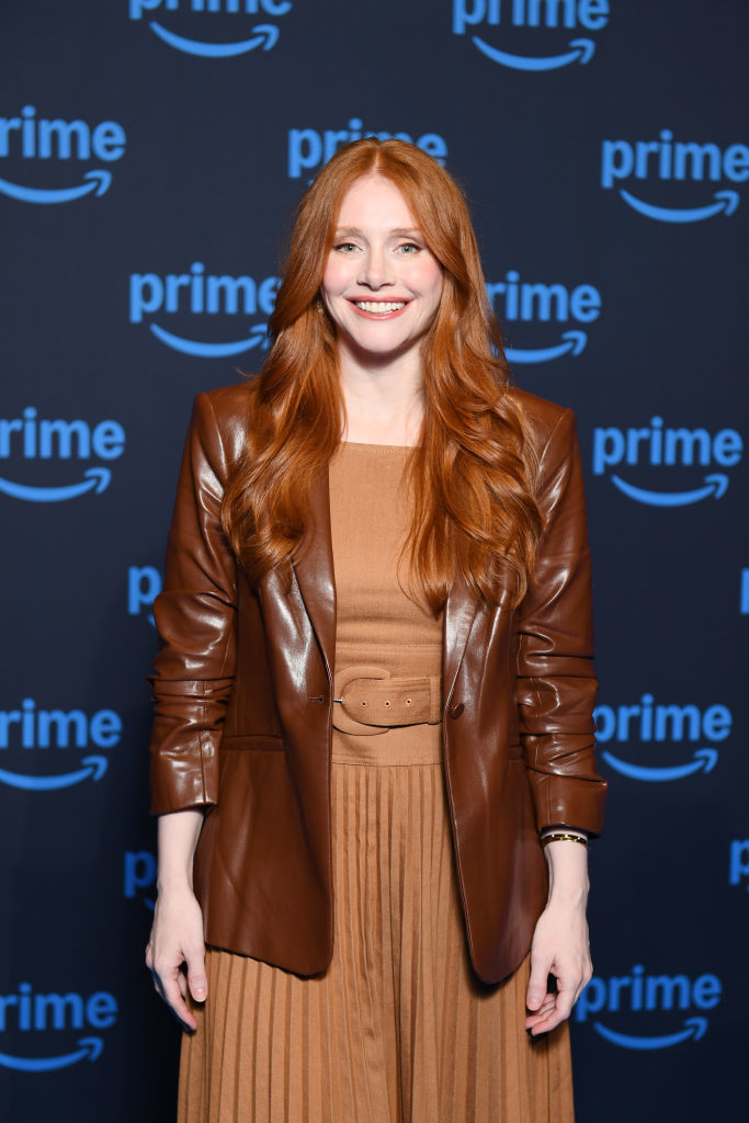 A person with long hair wears a leather blazer over a pleated dress, standing in front of a &quot;Prime&quot; branded backdrop at a style-related event