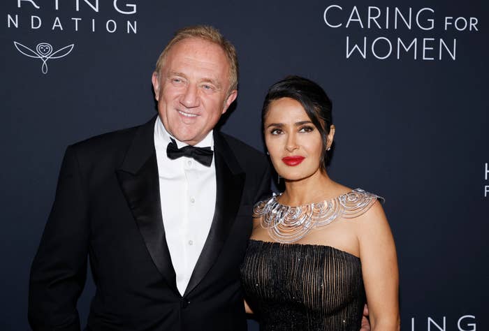 François-Henri Pinault, and Salma Hayek posing on a red carpet. Salma wears an elegant dark gown with metallic accents, and François-Henri, is in a classic tuxedo