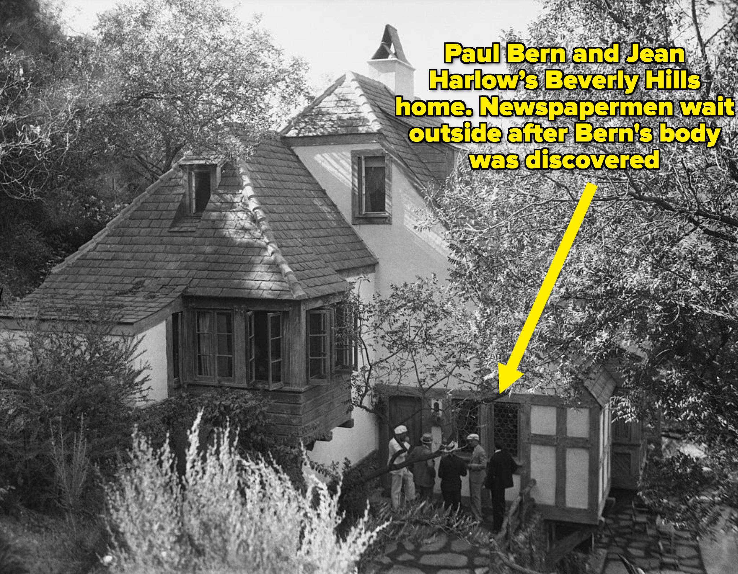 A vintage photo of a storybook-style house surrounded by trees. Three people stand near the entrance