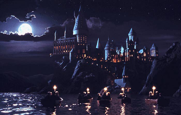 Boats with people approach a large, illuminated castle at night under a full moon, reminiscent of scenes from a fantasy film