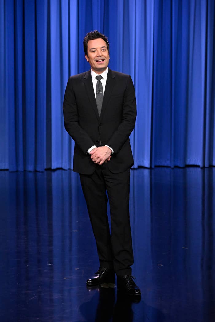 A man in a suit stands on a stage with a blue curtain background