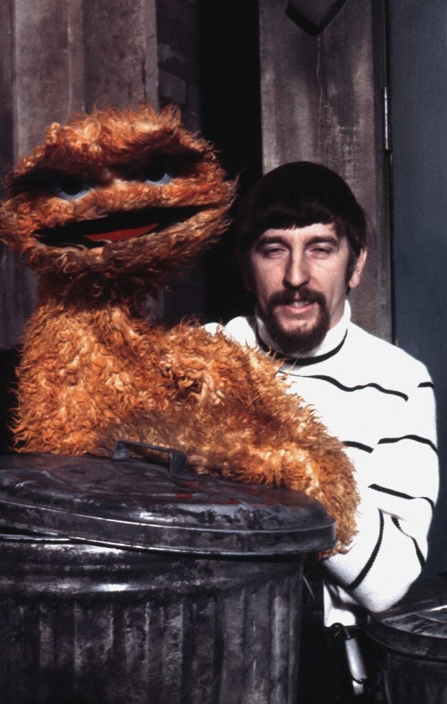 A person with a beard and a striped sweater poses with Oscar the Grouch from Sesame Street behind a trash can