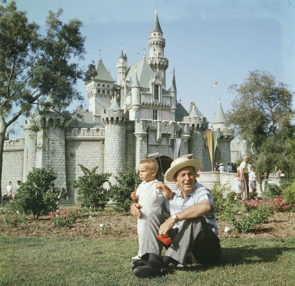 Man with a hat sits on grass with a child, in front of Sleeping Beauty Castle at Disneyland