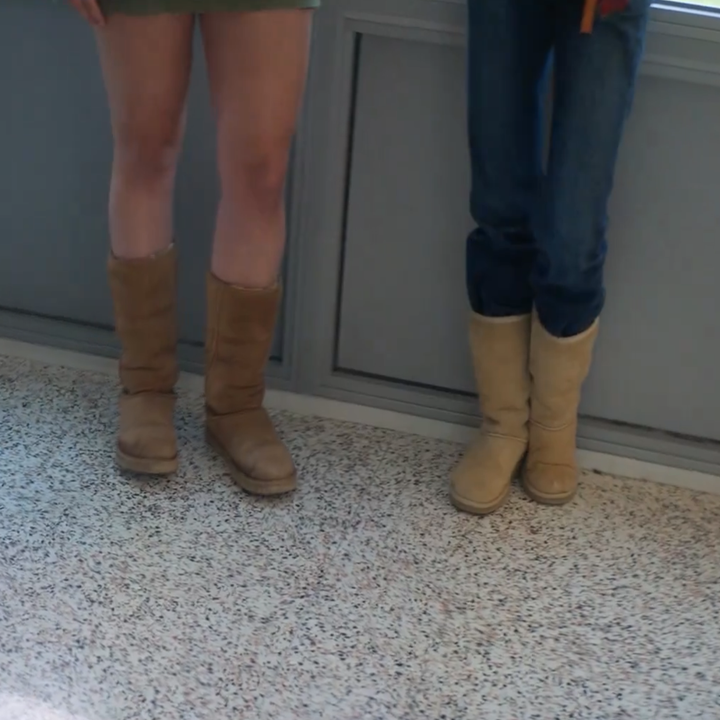 Two people stand indoors, showing their legs and boots. One wears a skirt, the other wears jeans