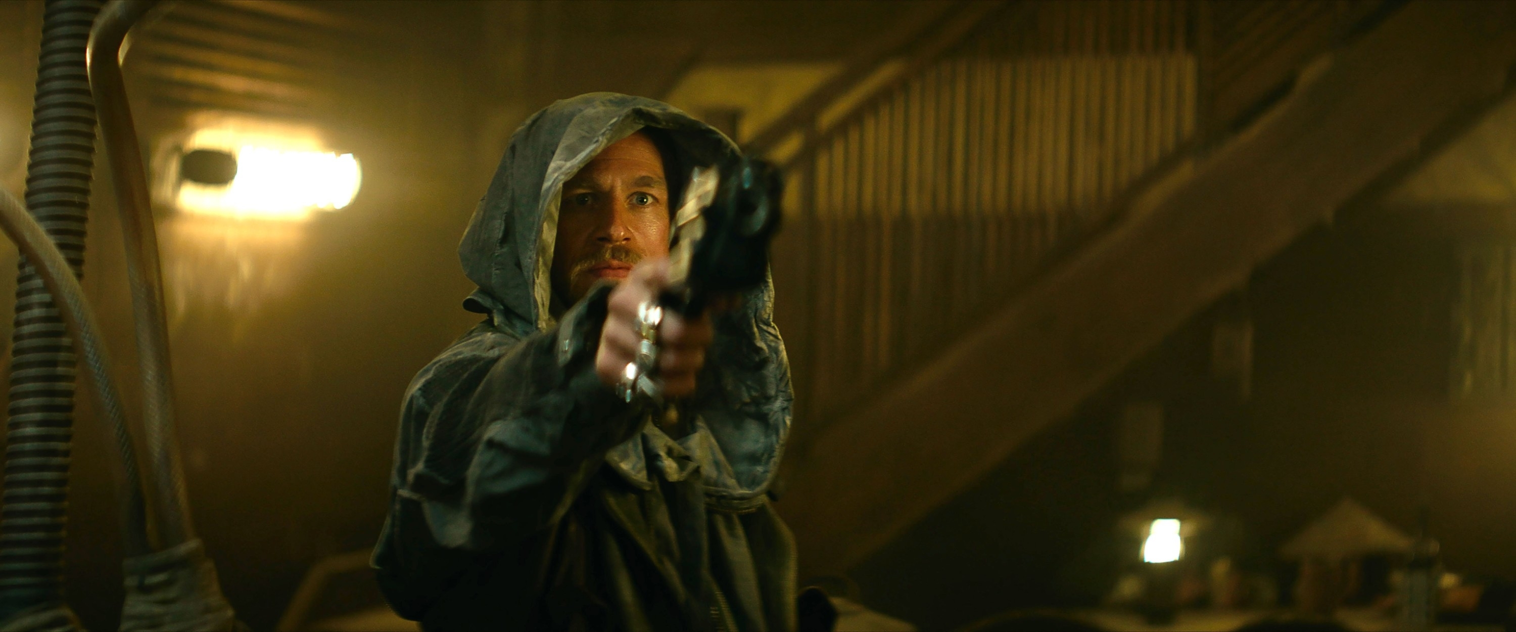 Charlie in a hooded cloak points a gun in a dimly lit, industrial setting