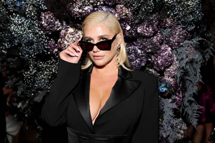 Kesha wearing sunglasses and a dark jacket, posing in front of a floral backdrop
