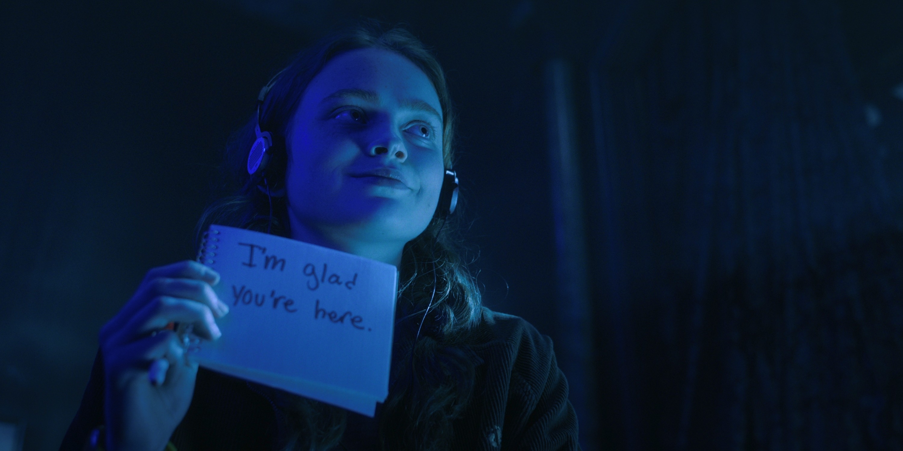 Sadie wearing headphones holds a small notebook with the handwritten message: "I'm glad you're here." Background is dimly lit