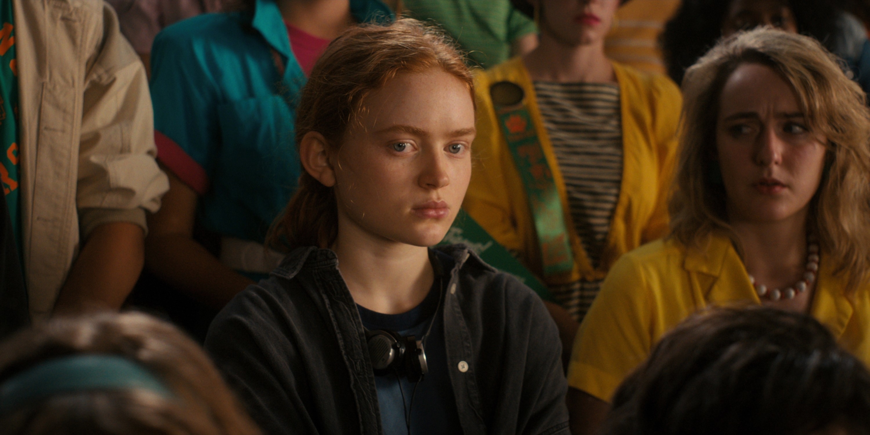 Sadie sits among a crowd, looking serious, in a scene. Others appear engaged in the background