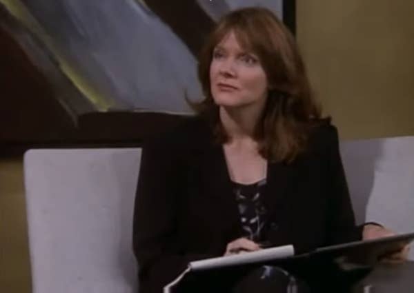 Maggie Baird in a blazer sits on a sofa, holding a folder, with abstract art in the background.
